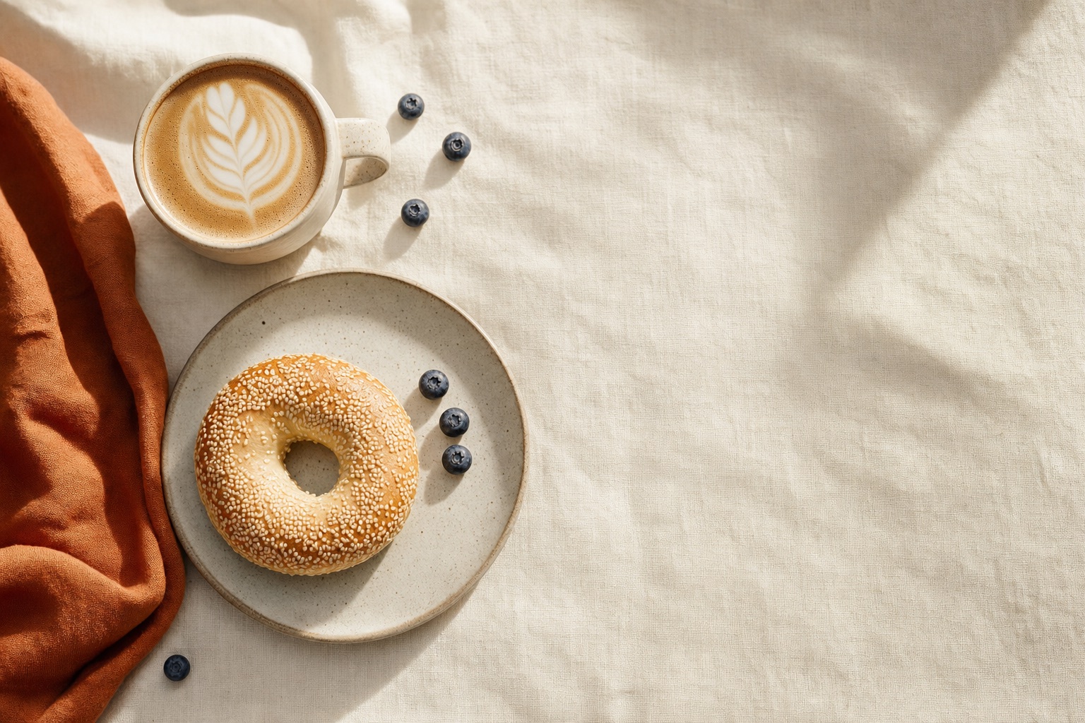 A latte and sesame bagel on cream linen in soft morning light